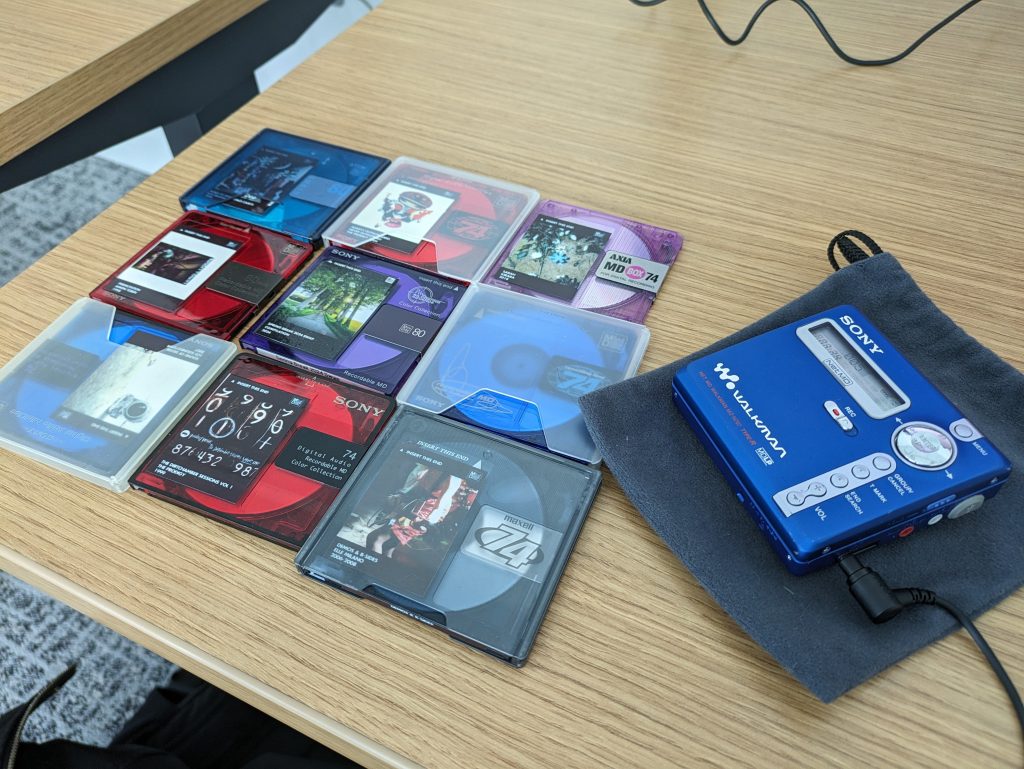 A collection of MiniDiscs with labels next to a blue Sony MZ-N707 player on top of a wooden desk.