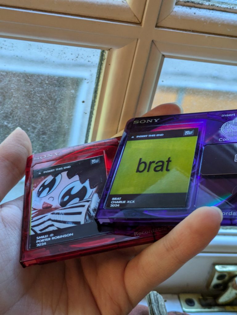 Two MiniDiscs with labels sitting next to a window. One is Brat by Charli XCX and the other is Smile by Porter Robinson.