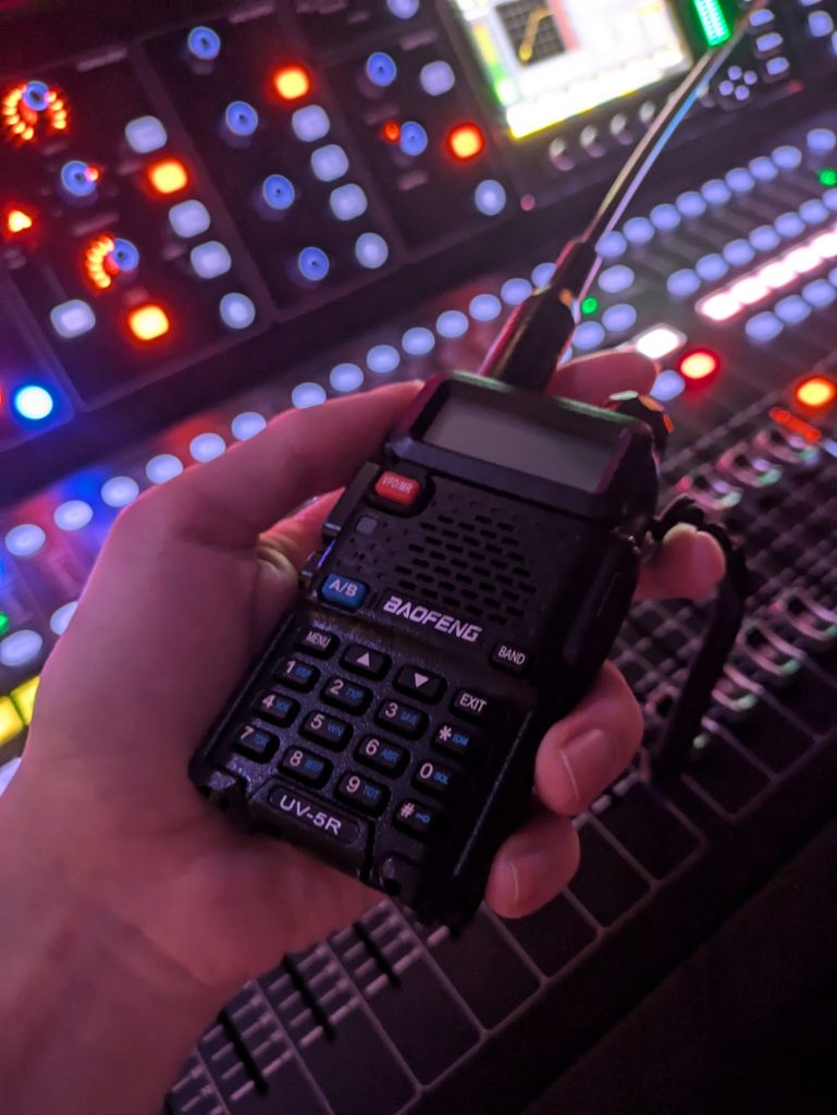 Riley's Baofeng handheld radio in front of a digital mixing console in a music venue.