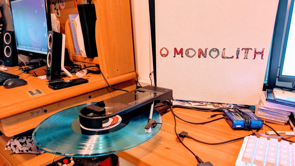 The Audio-Technica Sound Burger in action, playing Monolith by Squid. The record hangs off the side of a long rectangular device with a tonearm jutting out and sitting on top of the record. 