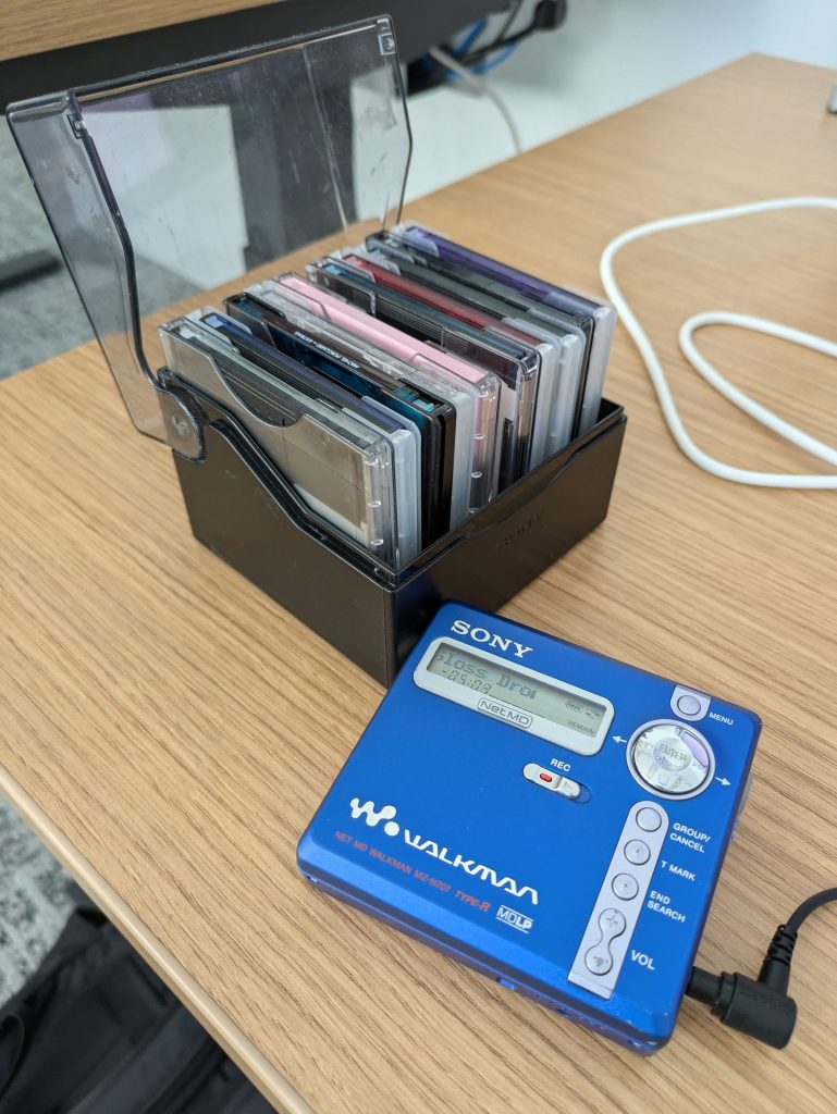 The Sony MZ-N707 sitting on a desk next to an open carrying case of MiniDiscs. 